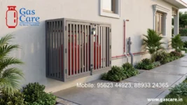 Gas Care's wall-mounted grey dual-cylinder LPG gas storage cage with closed slatted doors and padlock on a stucco house wall. Visible red cylinders inside. Features Gas Care By Stove-Technica logo. Neat red and white pipeline installation. Surrounded by lush, landscaped tropical plants, dwarf palms, and a paved concrete pathway. Contact numbers 95623 44923 and 88933 24365, with the website www.gascare.in displayed.